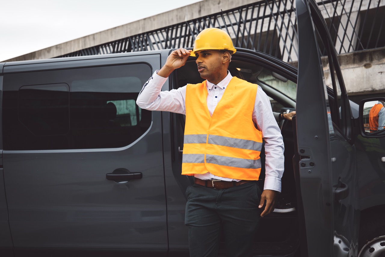 Construction worker in orange safety vest and yellow hard hat stands by an open door of a gray van.