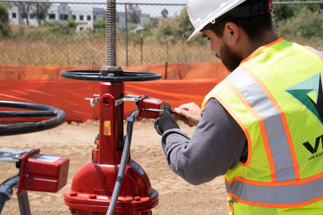 Worker in a hard hat and reflective vest inspects a red fire sprinkler valve outdoors.
