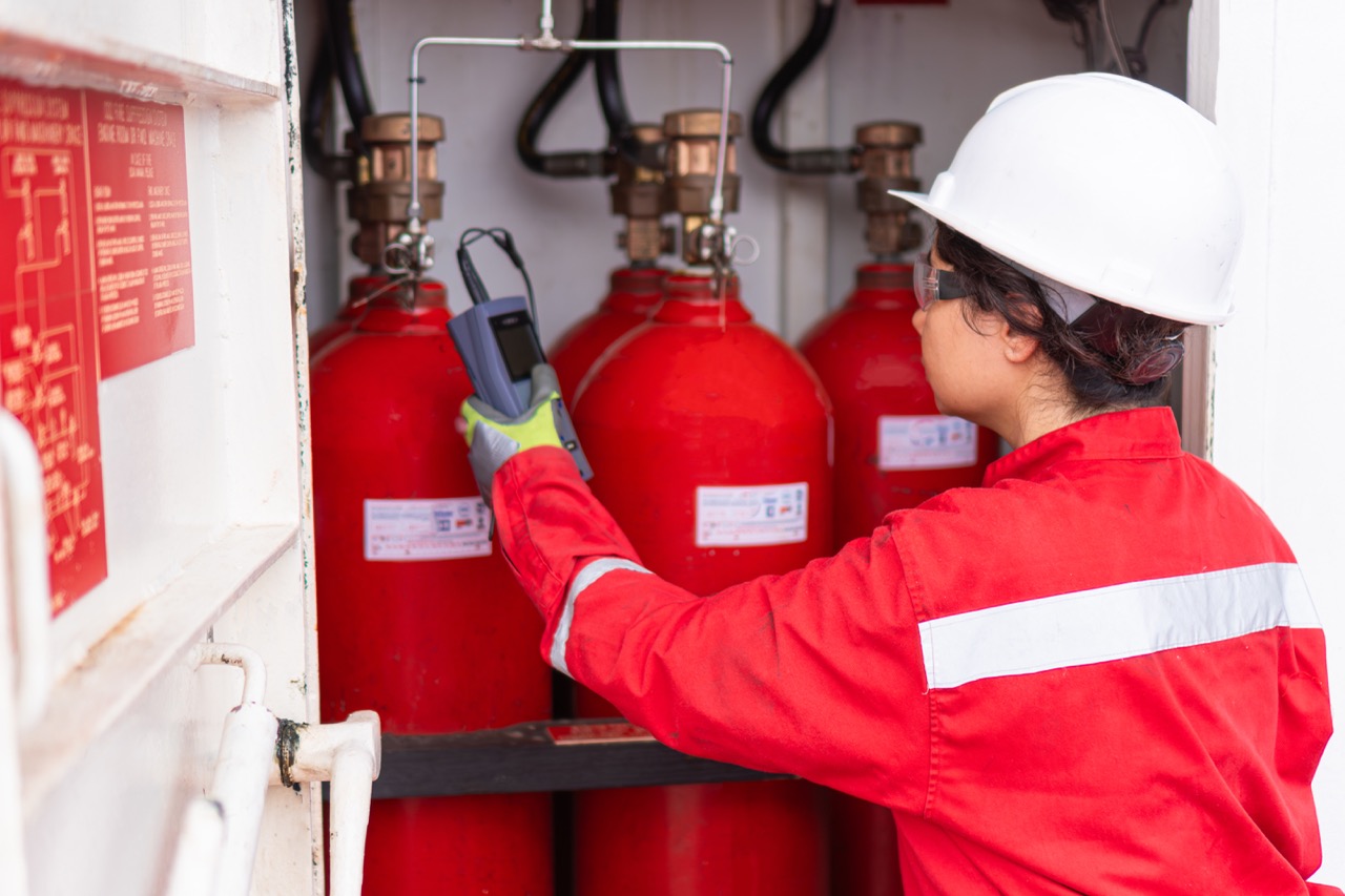 Worker in red uniform and white helmet inspects fire suppression system cylinders with a device.