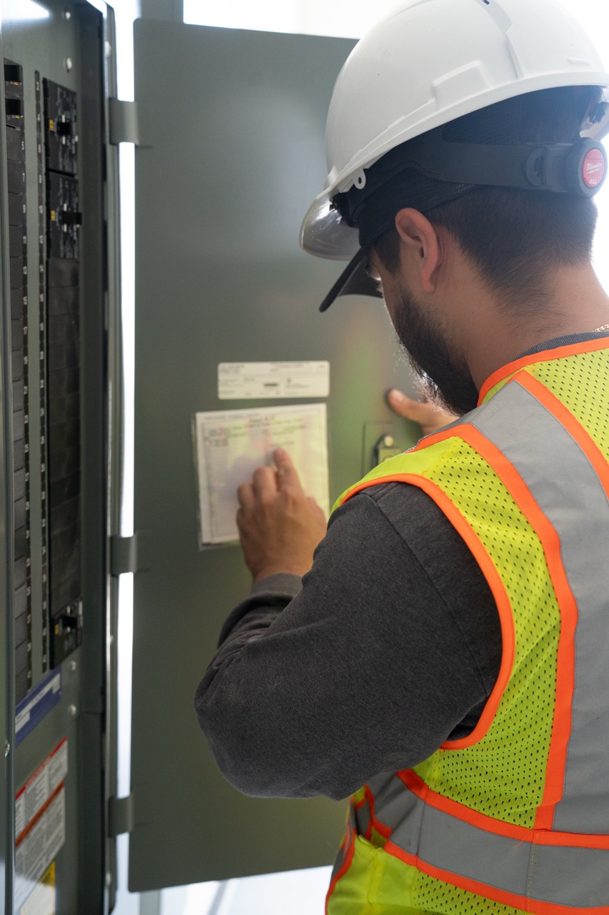 Worker in hard hat and reflective vest inspecting electrical panel with open circuit breaker box.
