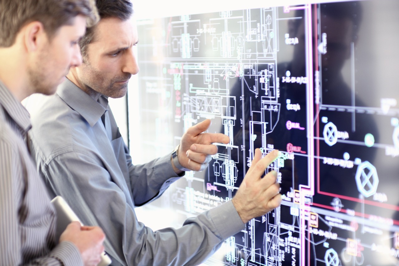 Two men discussing and pointing at a large digital architectural blueprint on a touchscreen display.