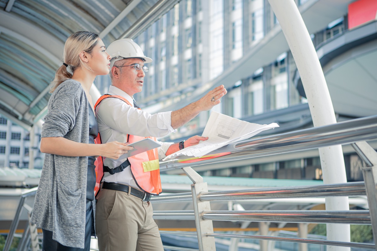Female and male architects discussing plans on site; man wears hard hat and safety vest.