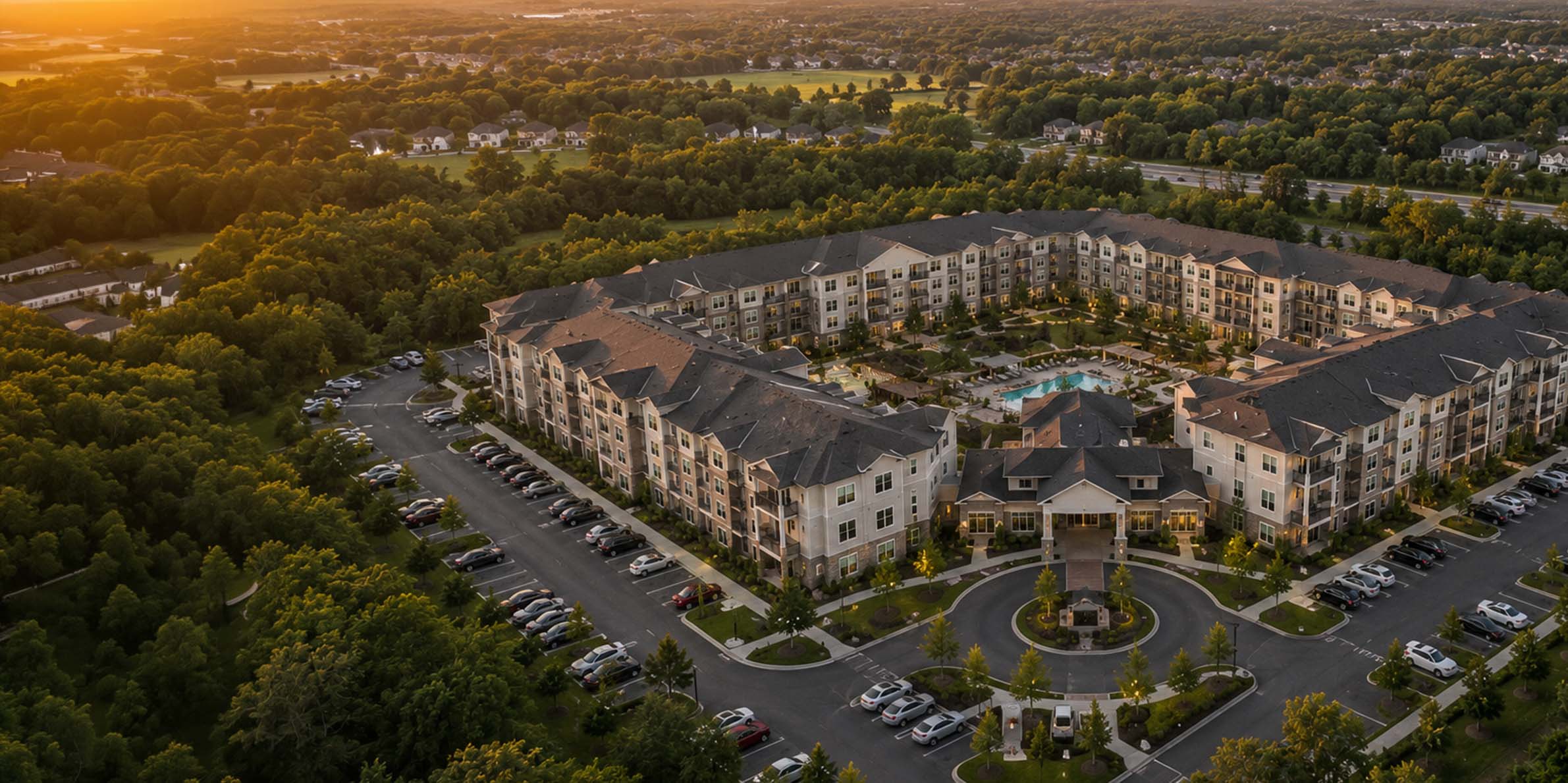 Aerial view of a large U-shaped apartment complex with a central pool and surrounded by greenery at sunset.