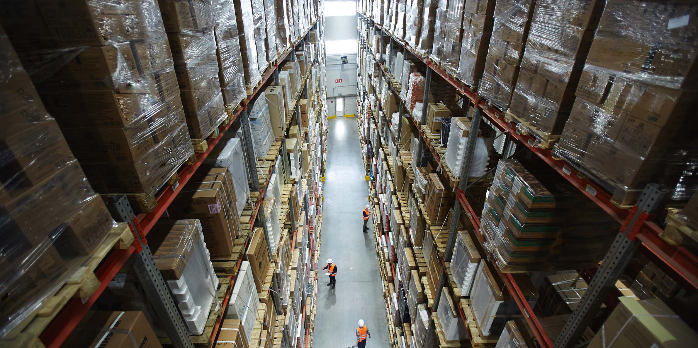 High-angle view of workers inspecting tall shelves stacked with boxes in a large warehouse aisle.