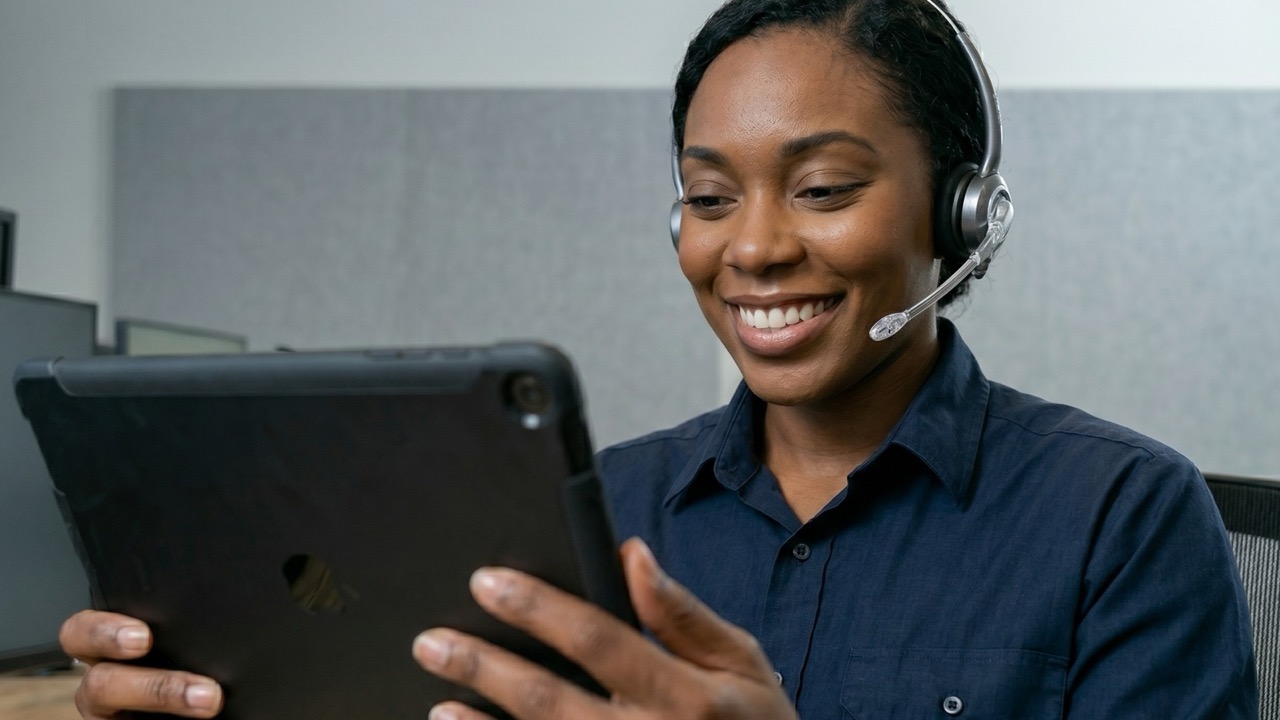 Smiling woman wearing headset, holding and looking at a tablet in an office setting.