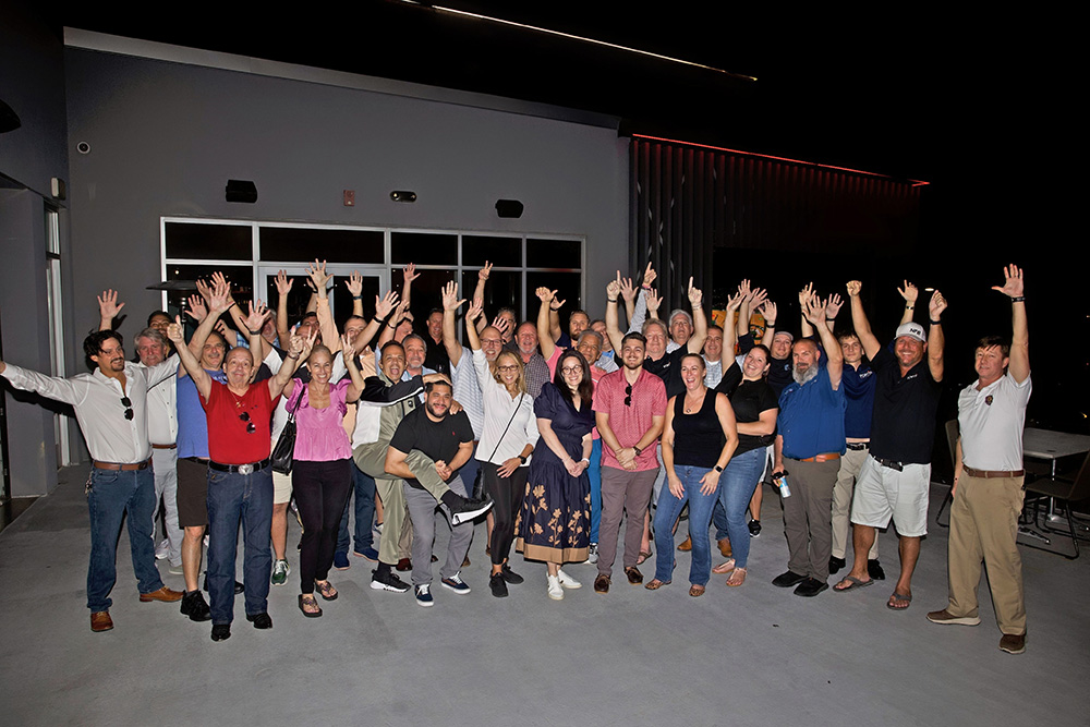 Large group of people outside at night with raised hands, smiling and posing for a photo.