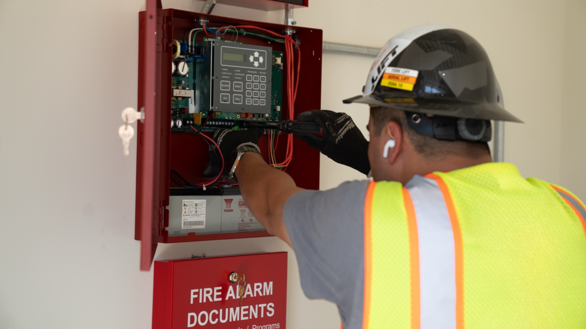 Technician in safety vest and helmet working on a fire alarm control panel inside a red box.