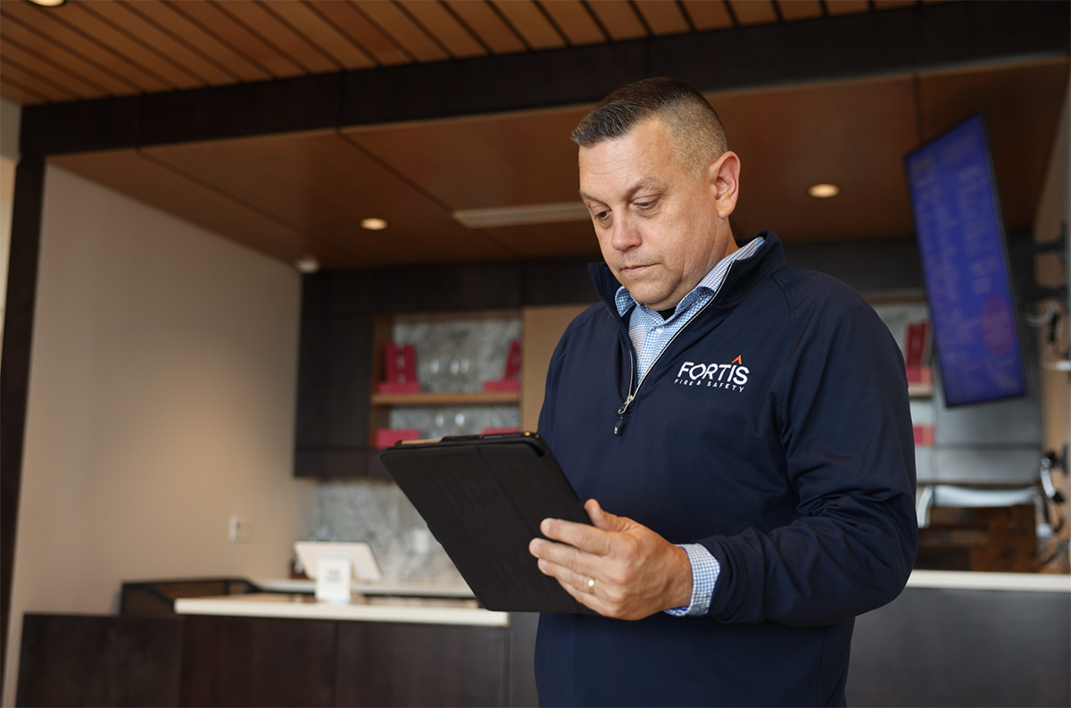 Man in navy blue Fortis Fire & Safety jacket holding and looking at a tablet in an office.