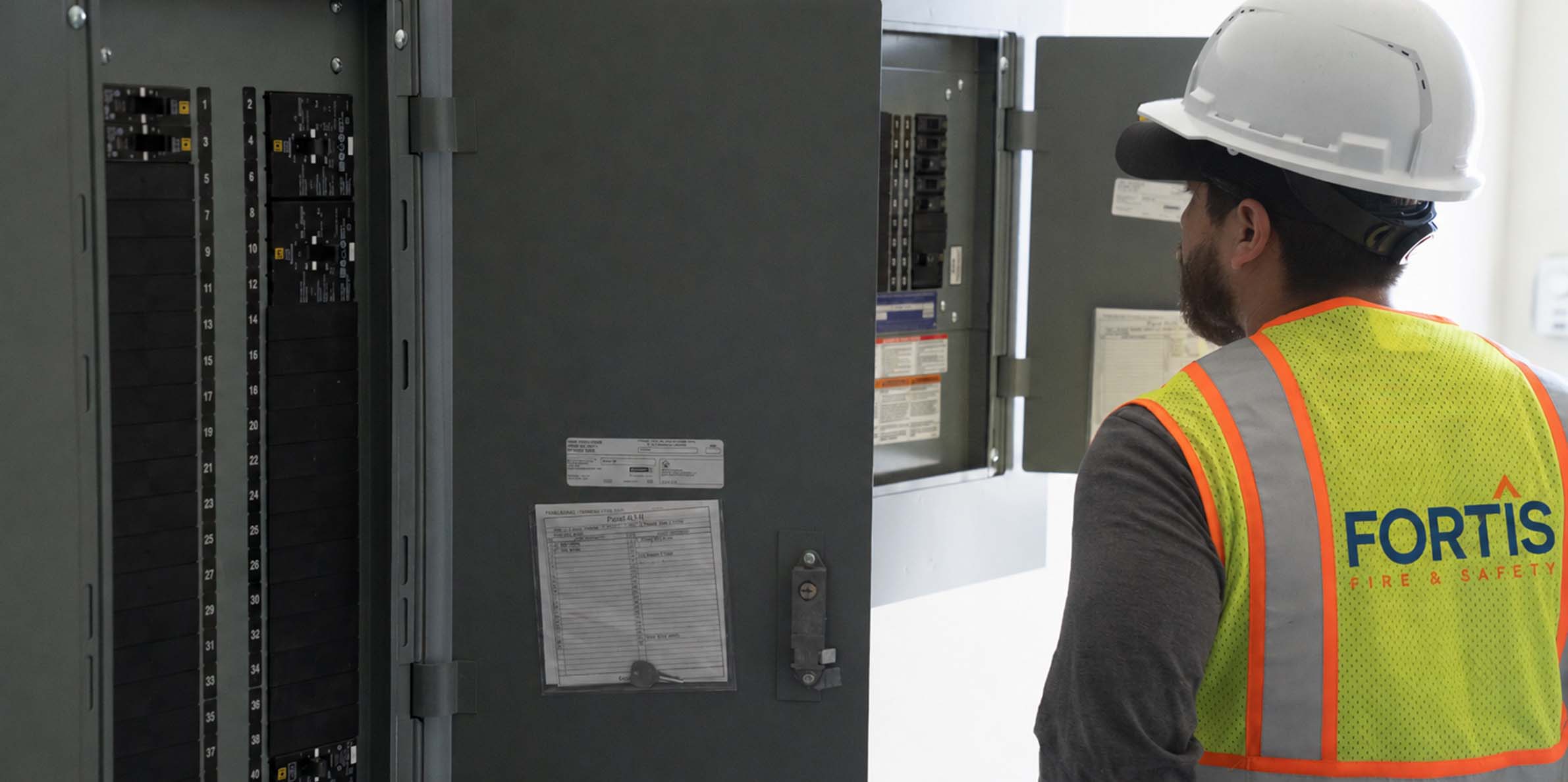 Worker in safety vest and helmet inspecting an electrical panel with open circuit breaker doors.
