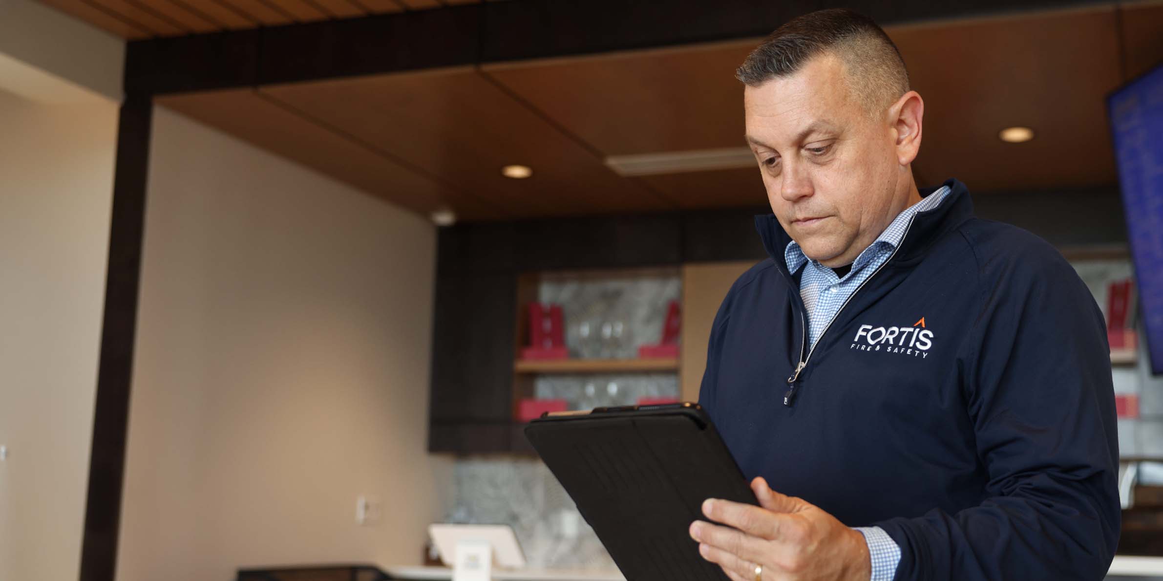 Man in navy Fortis Fire & Safety jacket looking at a black tablet in a modern office space.