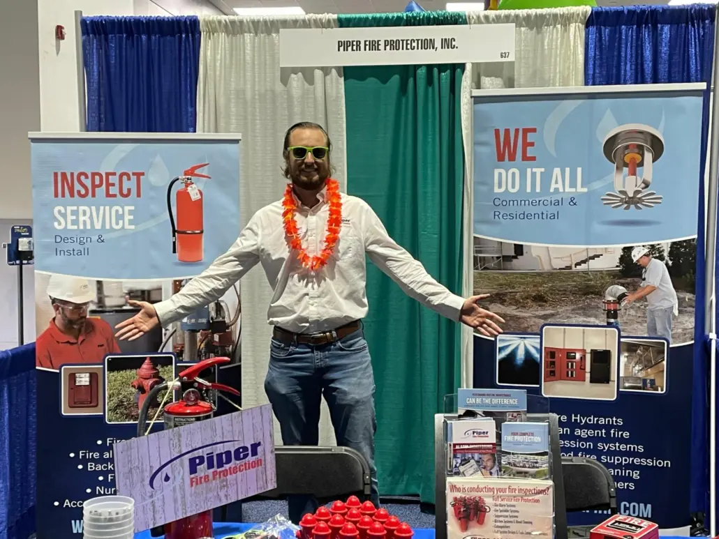 Man with sunglasses and orange lei at Piper Fire Protection booth with fire safety banners and products.