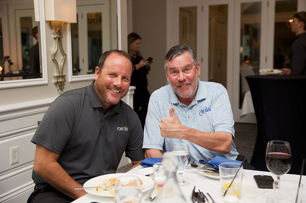 Two men sit at a table smiling; one gives a thumbs up, both wearing branded polo shirts at a dinner.