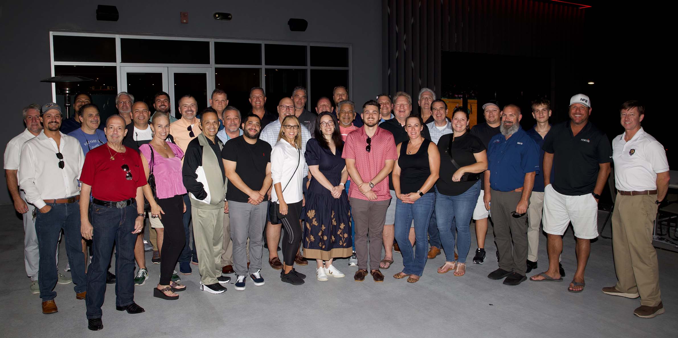 Group of 30 adults posing together outside a building at night, smiling at the camera.