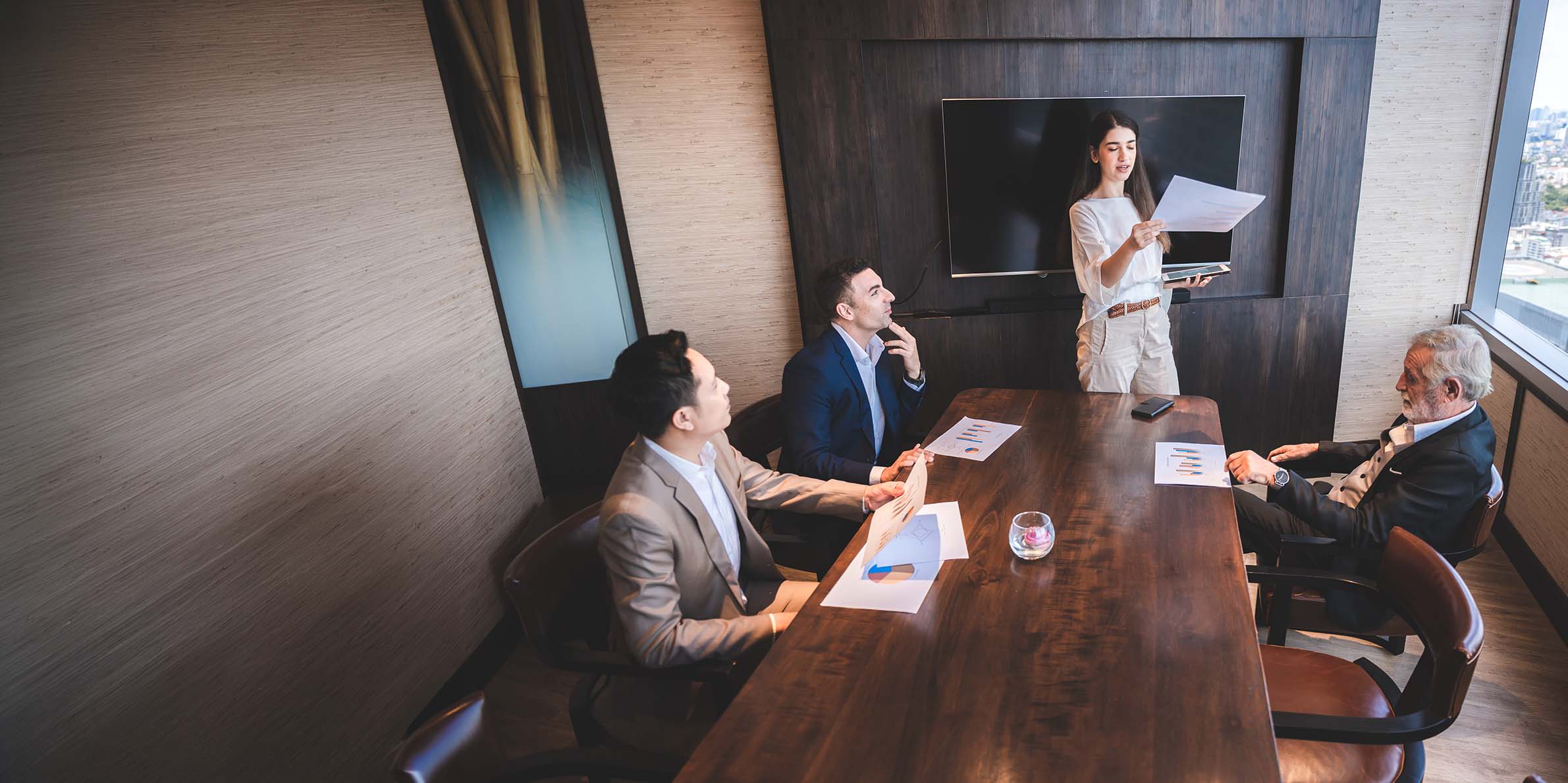 Four businesspeople in a meeting room with documents, a woman stands presenting to three seated men.