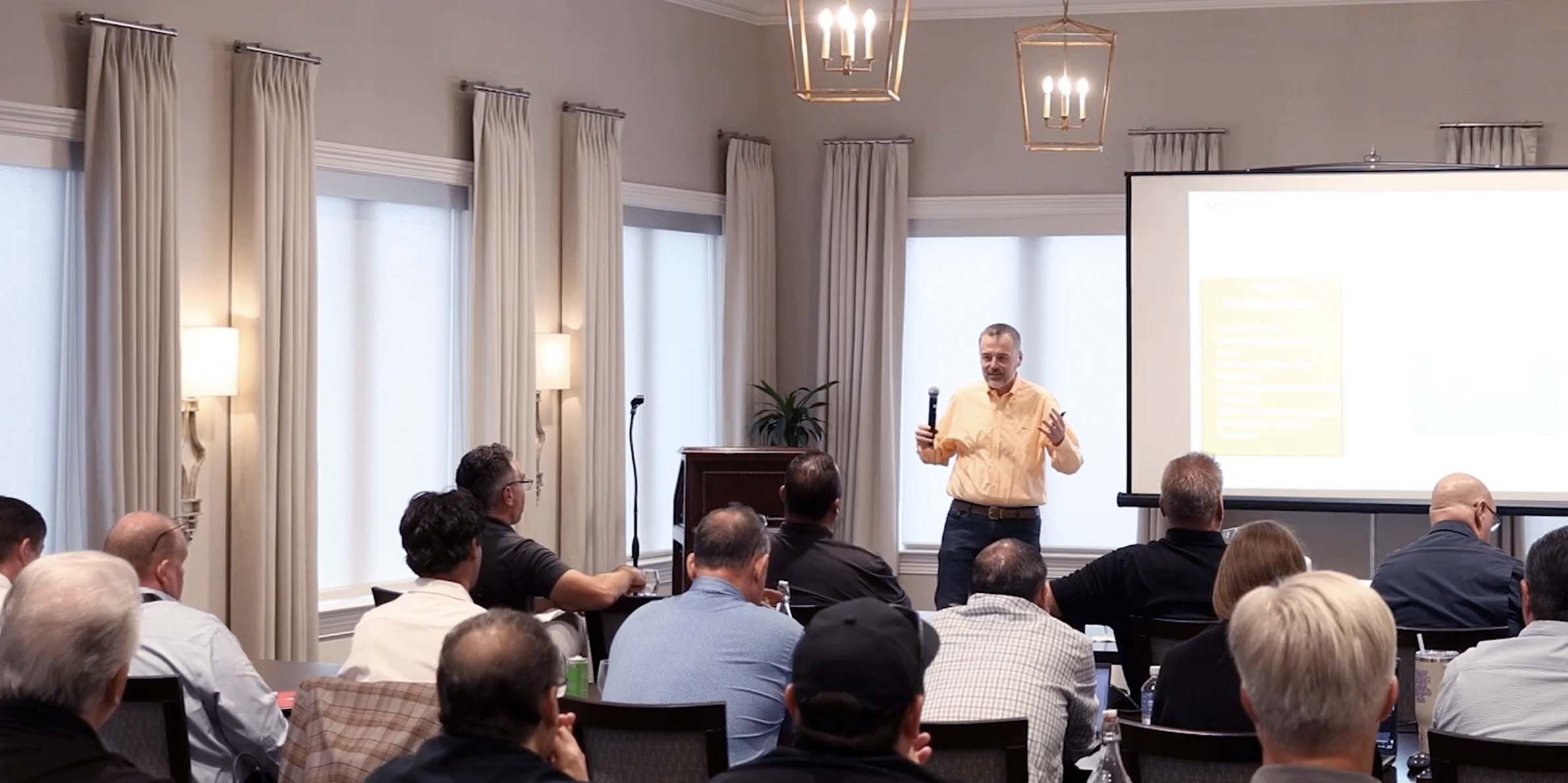 Man speaking with microphone to seated audience in a conference room with a projector screen.