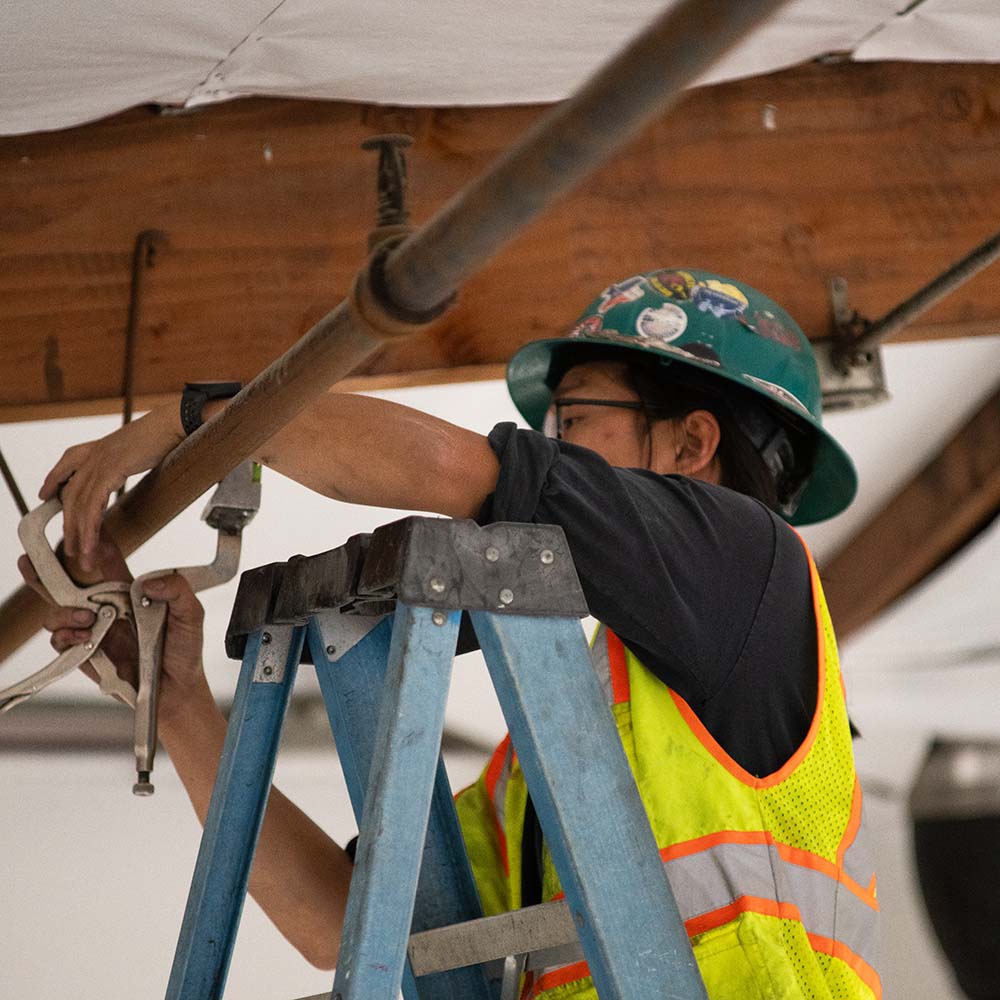Worker wearing a green helmet and safety vest fixing a pipe overhead on a ladder.