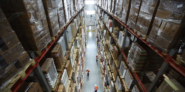 Warehouse aisle with tall shelves stacked with boxes and two workers wearing orange safety vests.