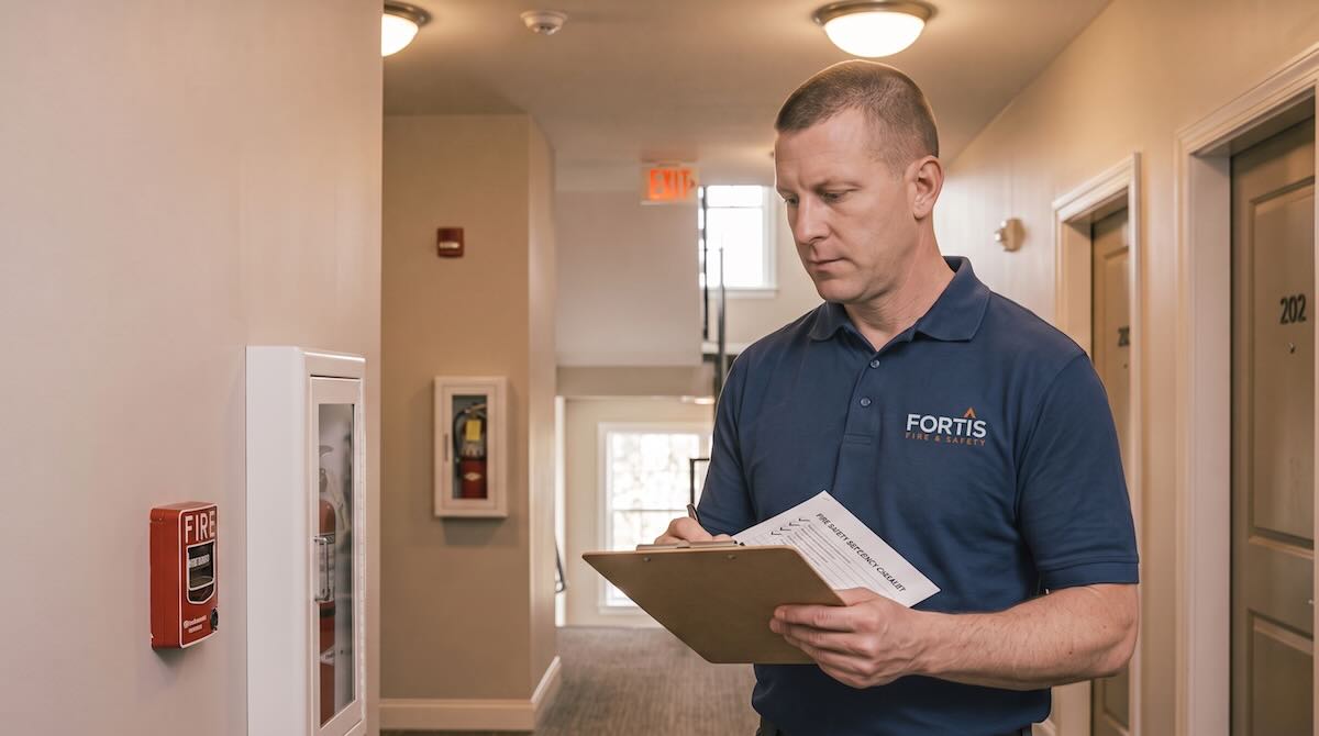 Man in Fortis uniform checking fire safety equipment and filling a checklist on a clipboard in a hallway.