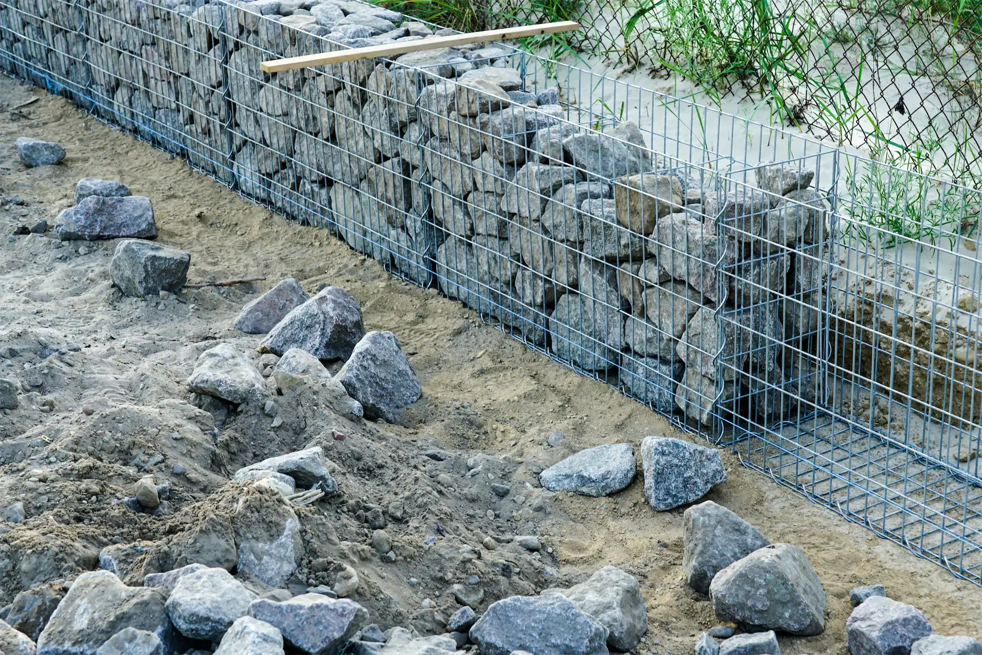 Mur de soutènement en gabions en construction avec des pierres empilées dans des cages métalliques sur un sol de terre.
