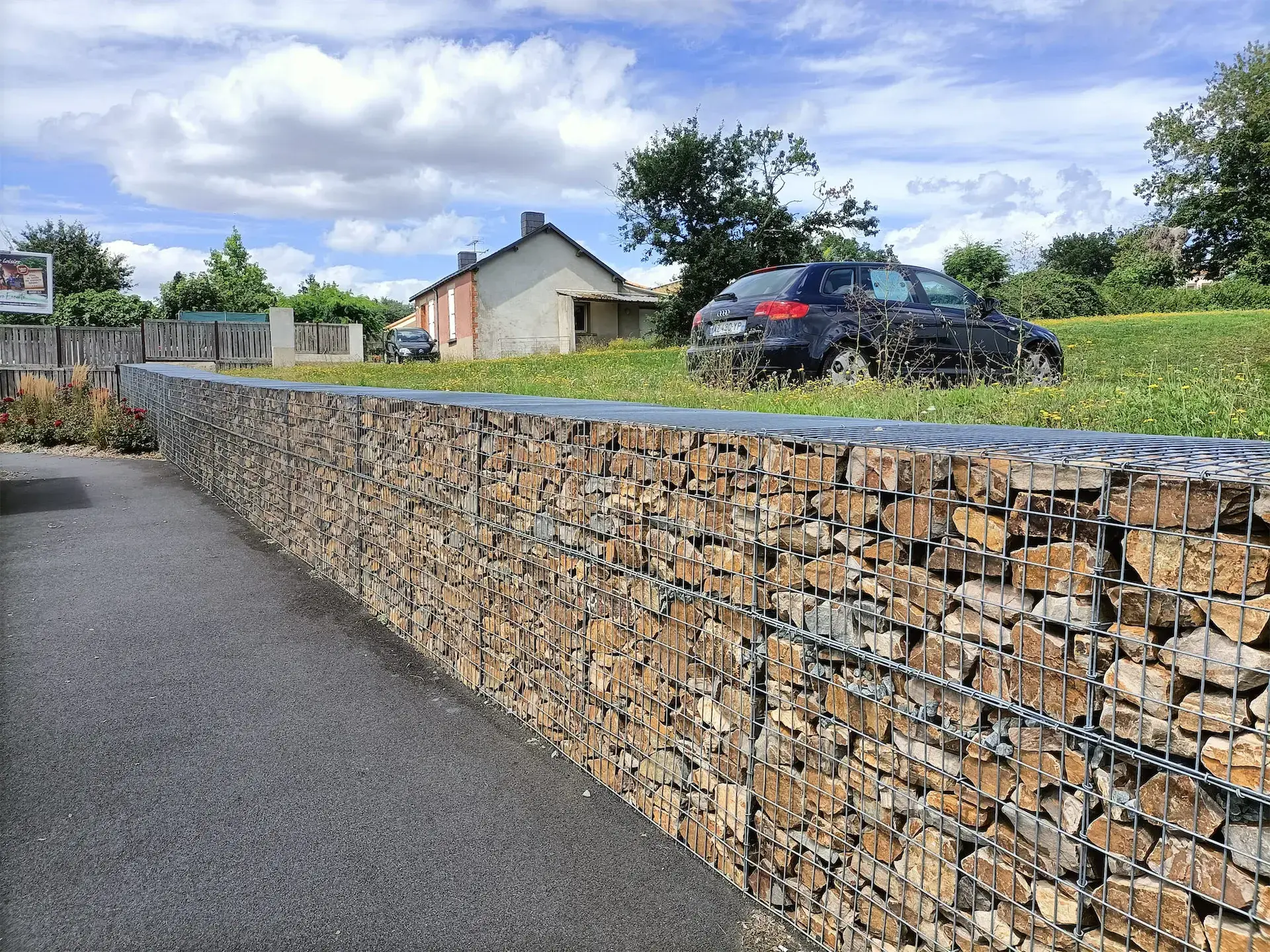 Mur gabion en pierres avec une voiture noire garée sur une pelouse à côté d'une maison sous un ciel partiellement nuageux.