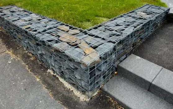 Mur de soutènement en gabion rempli de pierres grises formant un angle, adjacent à des escaliers en béton.