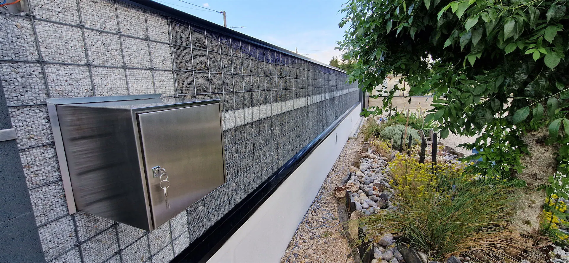 Boîte en acier inoxydable verrouillée fixée à un mur en gabions près d'un jardin avec des plantes et des rochers.
