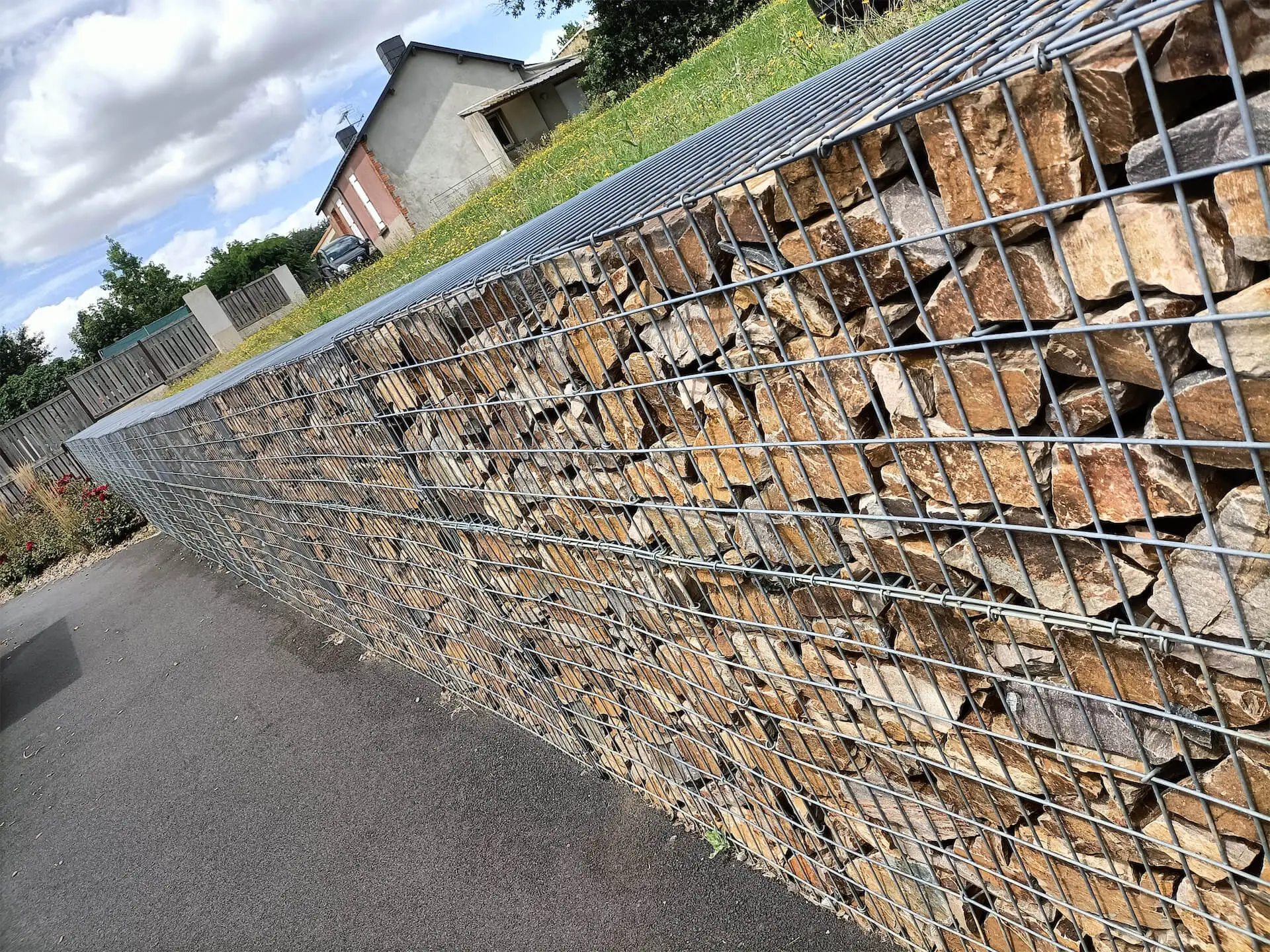 Une barrière gabion remplie de pierres marron le long d'un trottoir, avec des maisons et de l'herbe à l'arrière-plan.