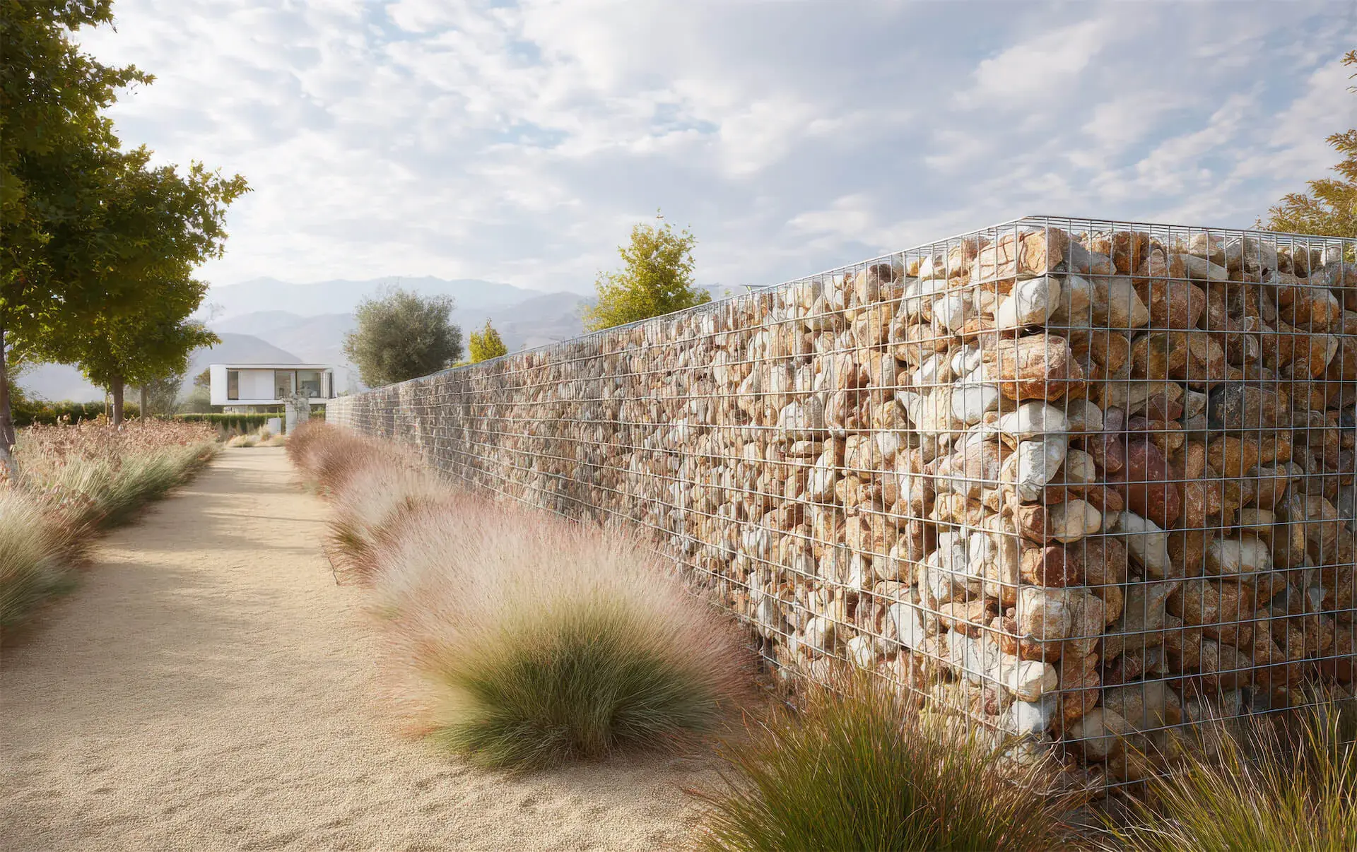 Chemin de terre bordé d'herbes décoratives et d'un mur en gabion rempli de pierres, avec une maison moderne en arrière-plan sous un ciel partiellement nuageux.