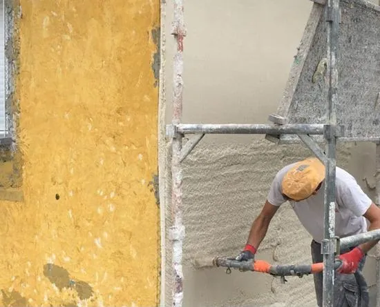 Un ouvrier applique du crépi gris clair sur un mur à côté d'une façade jaune, en utilisant un tuyau sous un échafaudage.