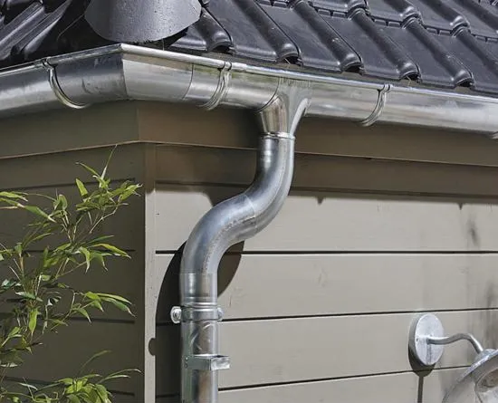 Gouttière en métal argenté incurvée fixée sur l'angle d'un bâtiment avec un toit à tuiles noires.