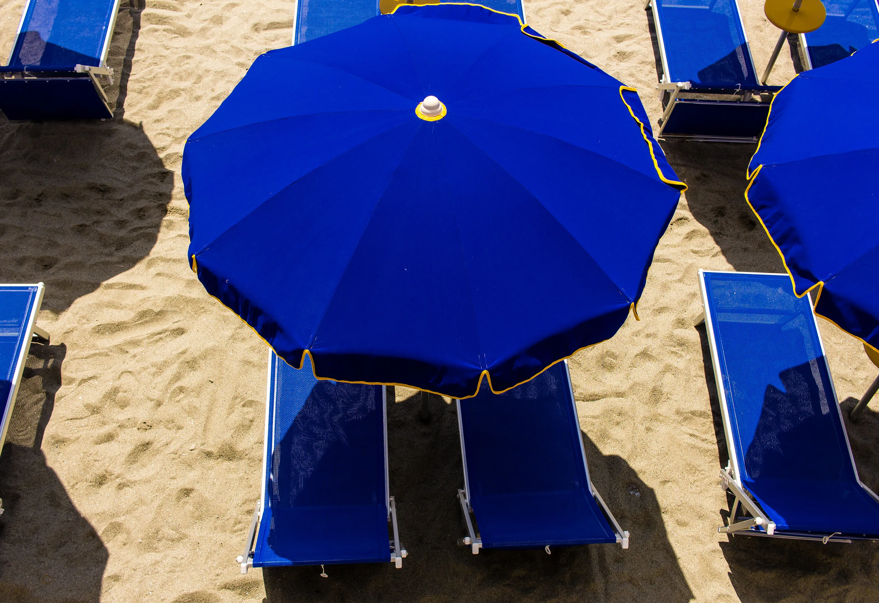 Zwei blaue Liegestühle unter einem blauen Strandsonnenschirm auf Sand.
