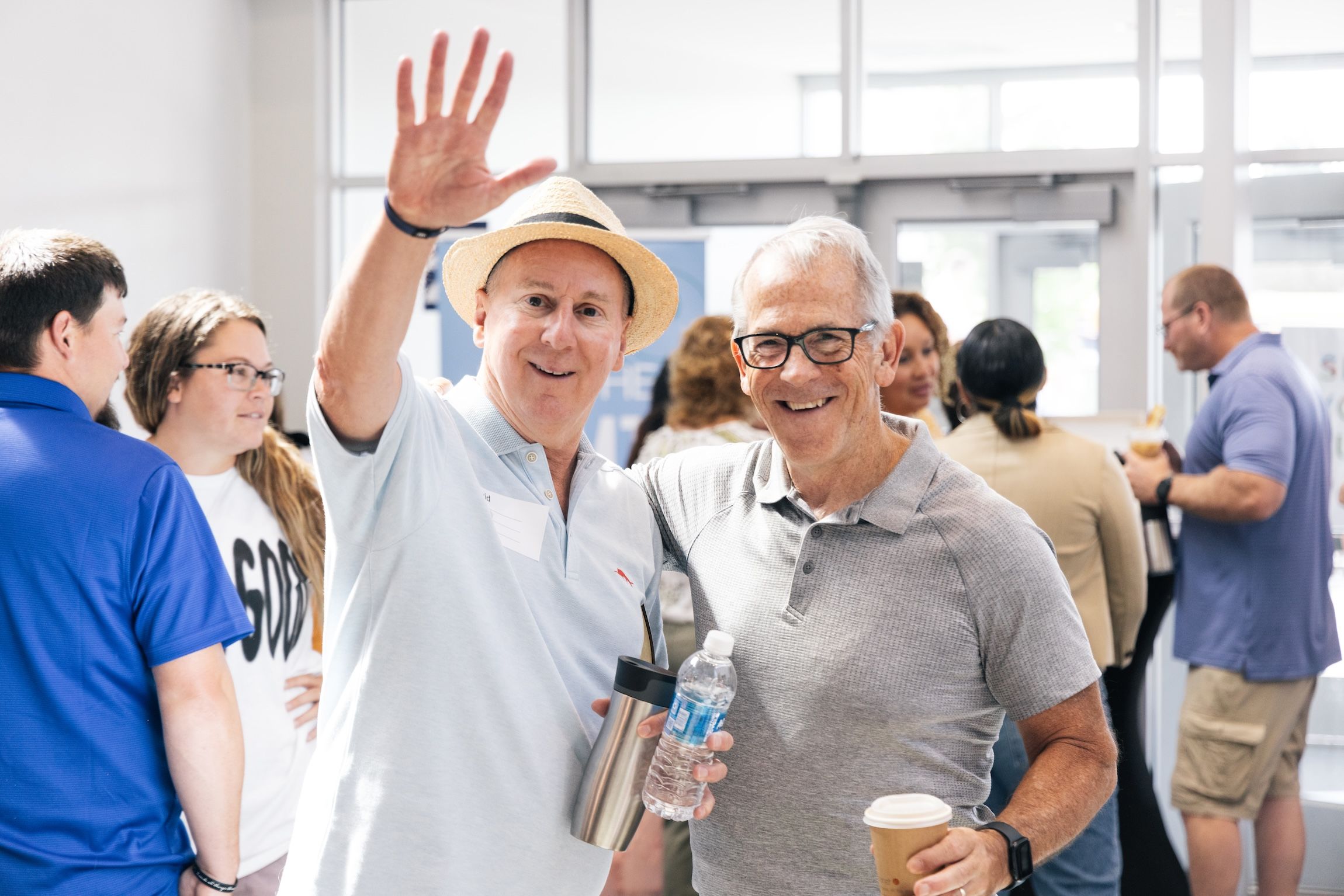 Two older men smiling, one wearing a hat and waving, holding a water bottle and a metal tumbler, the other holding a coffee cup, with people socializing in the background in a bright indoor setting.