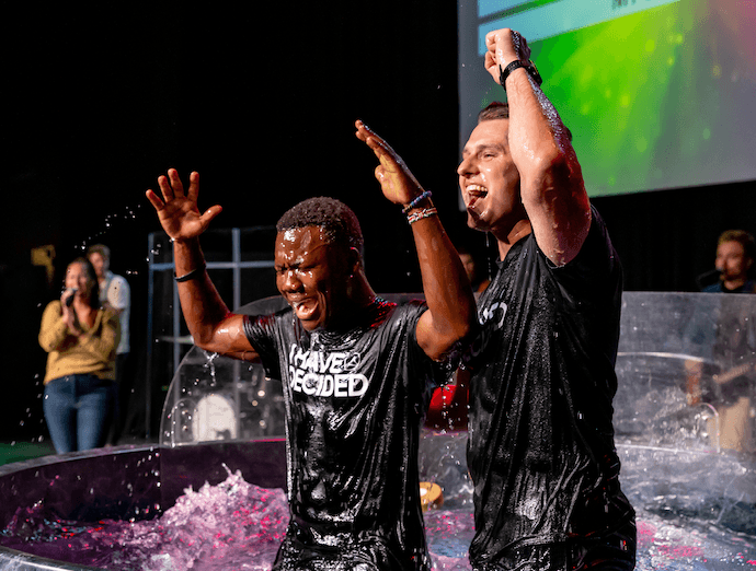 Two men in wet black t-shirts with one reading 'I HAVE DECIDED' raising their hands in celebration during a baptism ceremony.