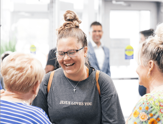 Smiling woman wearing glasses and a 'Jesus Loves You' shirt talking with two other women in a bright indoor setting.