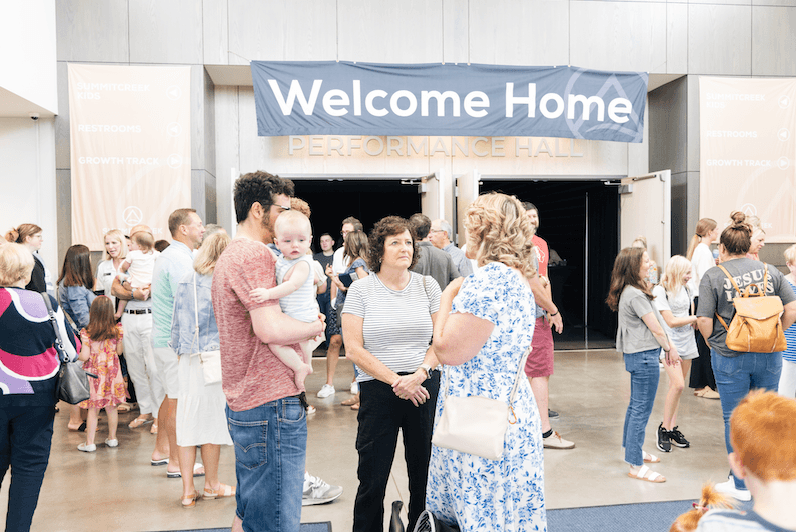 People of various ages, including a man holding a baby, gathered and socializing inside a building under a 'Welcome Home' banner above double doors labeled 'Performance Hall'.