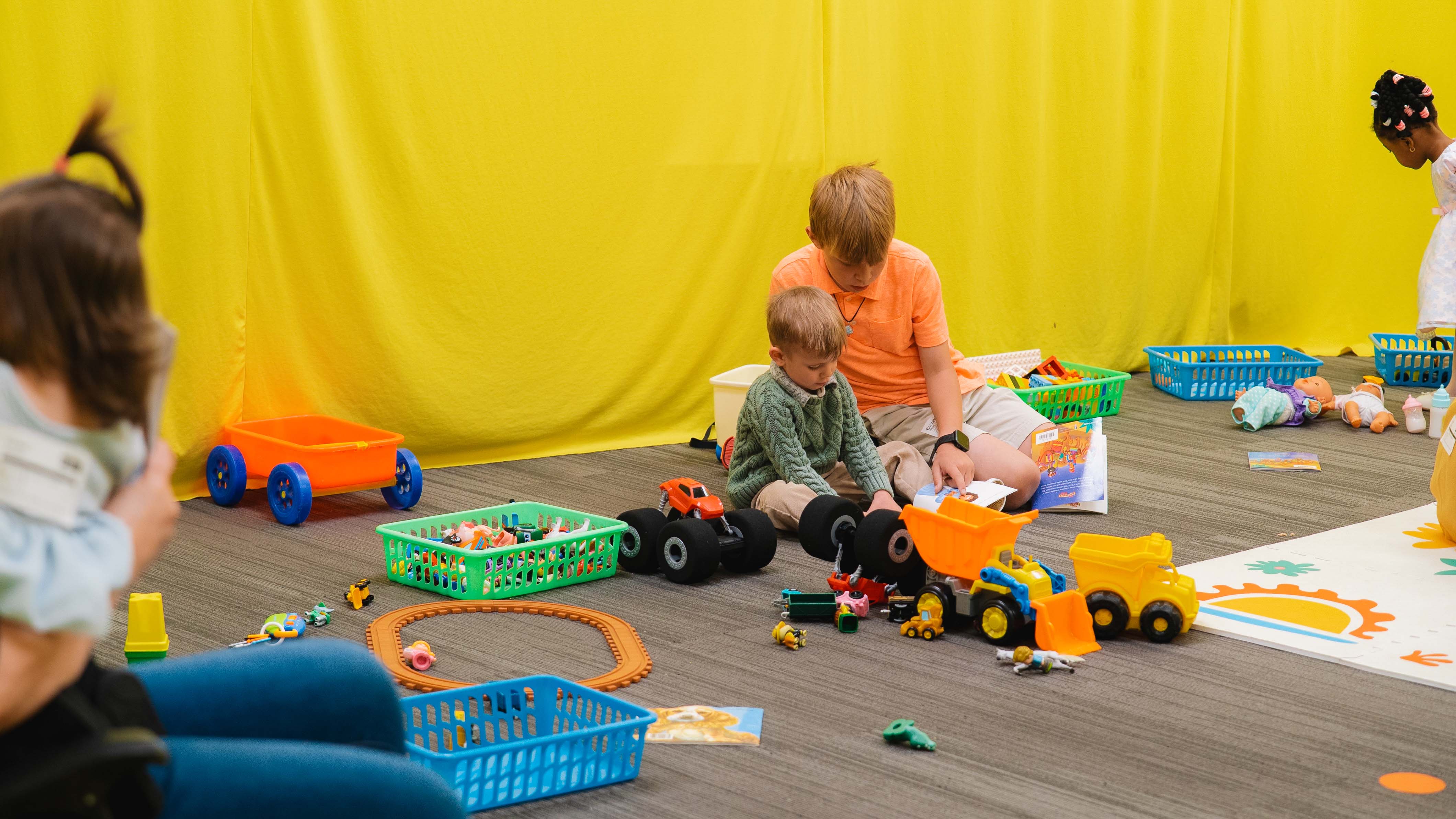 Two boys sitting on the floor with toys and books around them, playing and reading against a yellow background.