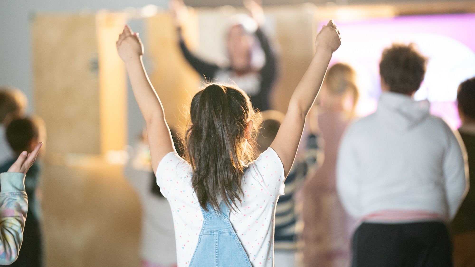 Child with a ponytail and denim overalls raising both arms in a bright indoor setting with a blurred group of people facing a speaker.