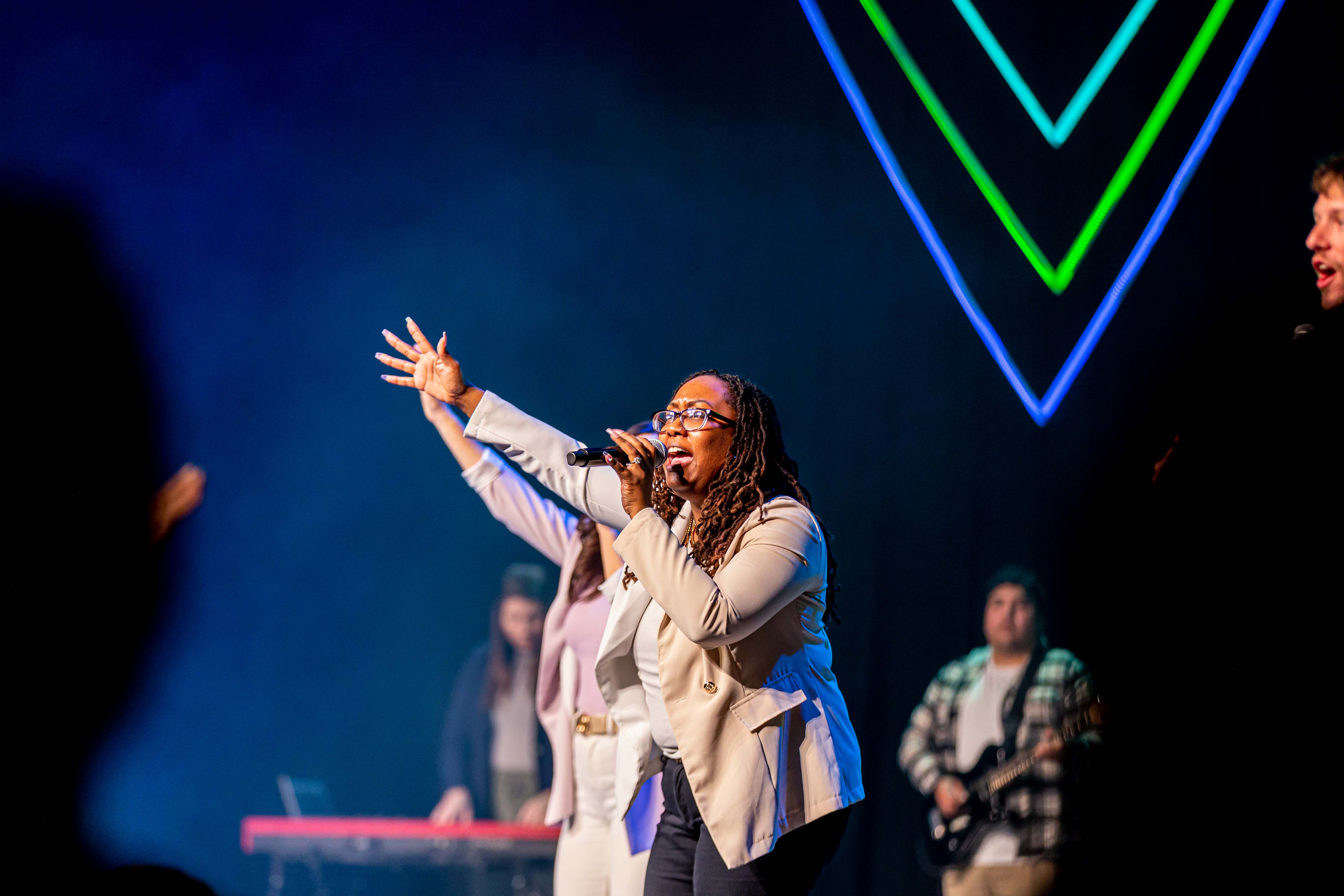 Woman with glasses passionately singing into a microphone on stage with band members and neon lights in background.