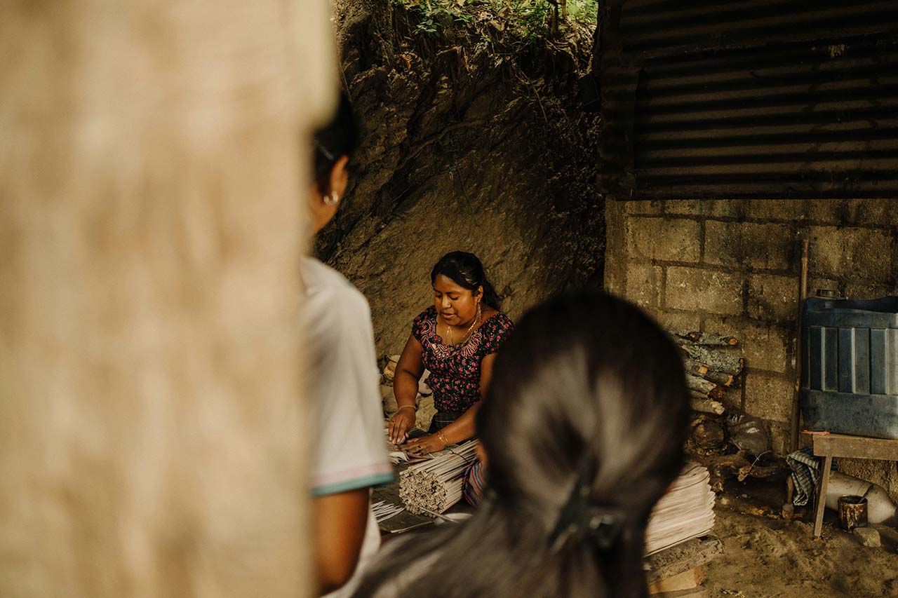 Woman weaving or working with reeds or sticks inside a rustic shelter with another person blurred in foreground.