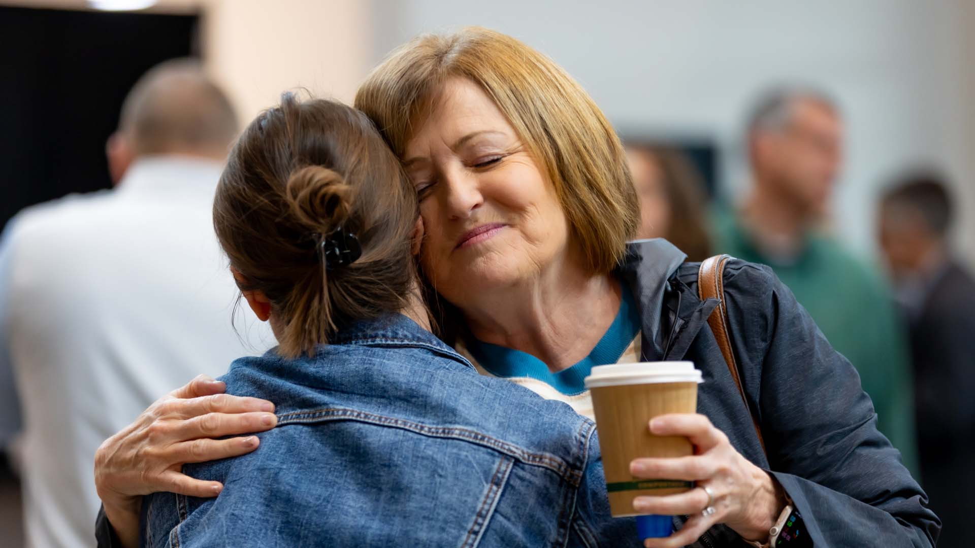 Two women warmly embracing, one holding a coffee cup and smiling with eyes closed.
