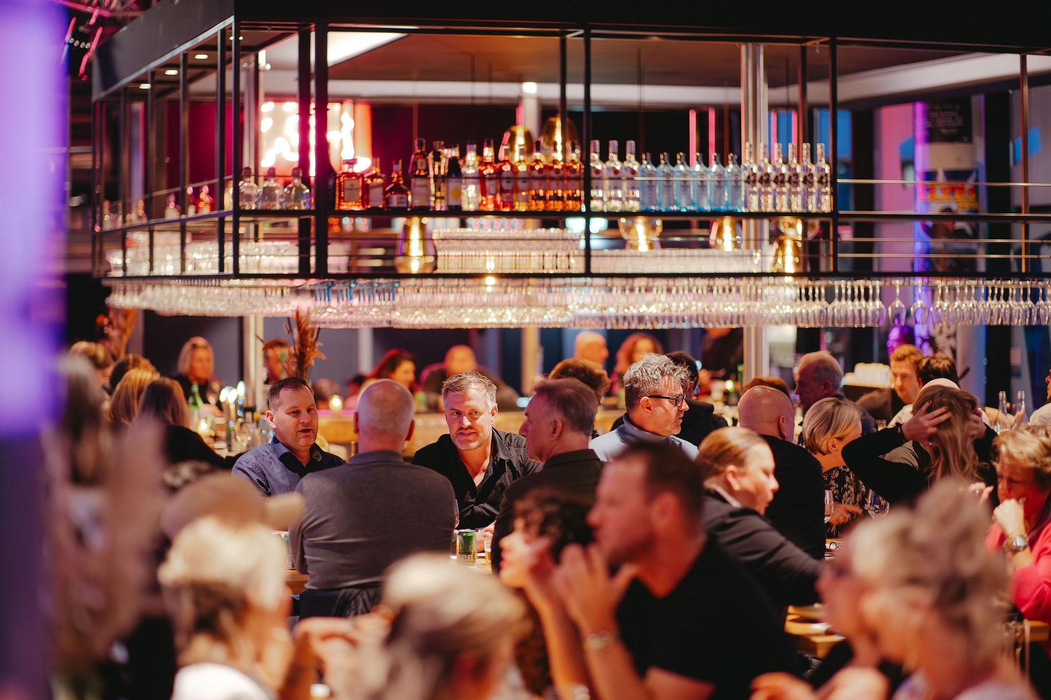 Gasten aan lange tafels onder de bar van BAUT Backstage