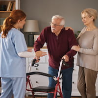 Elderly man using a walker supported by a woman and assisted by a nurse in a home setting.