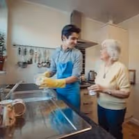Young woman wearing yellow gloves washing dishes in a kitchen with an elderly woman watching and smiling.
