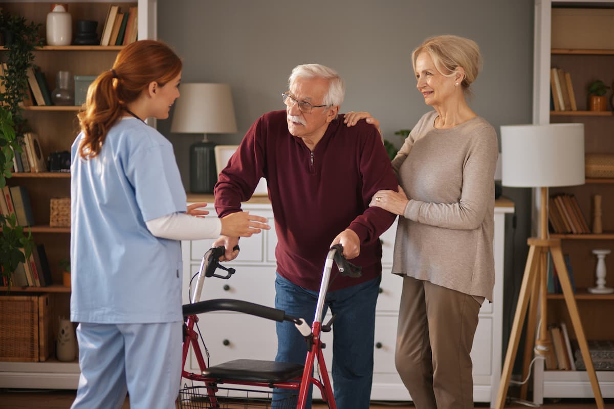 Elderly man using a walker supported by a smiling woman and a caregiver in blue scrubs in a cozy living room.