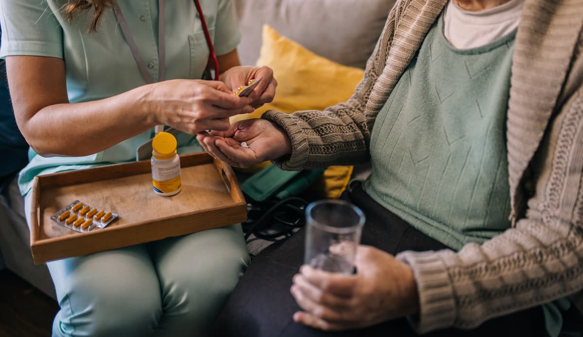 A healthcare worker giving pills to an elderly person sitting on a couch holding a glass of water.