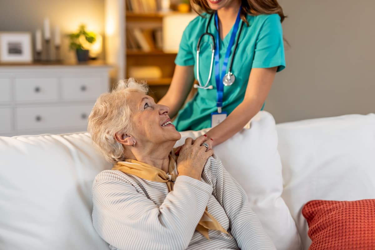 Elderly woman sitting on a white couch looking up and smiling at a smiling female healthcare worker in green scrubs with a stethoscope.