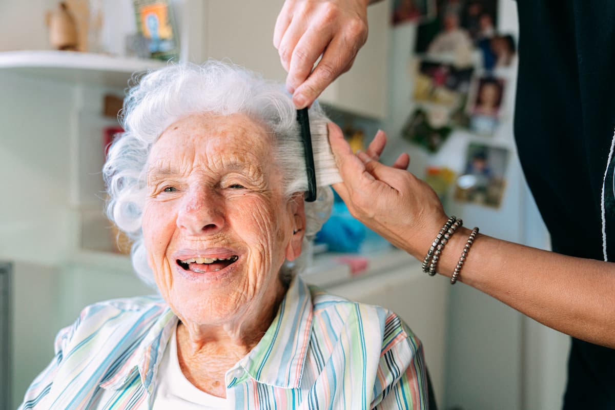 Elderly woman with white hair smiling while getting her hair combed by a person wearing bracelets.