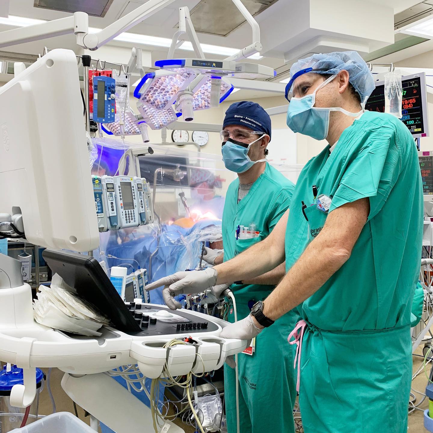 Two medical professionals in green scrubs, masks, and gloves operating medical equipment in a brightly lit hospital room.