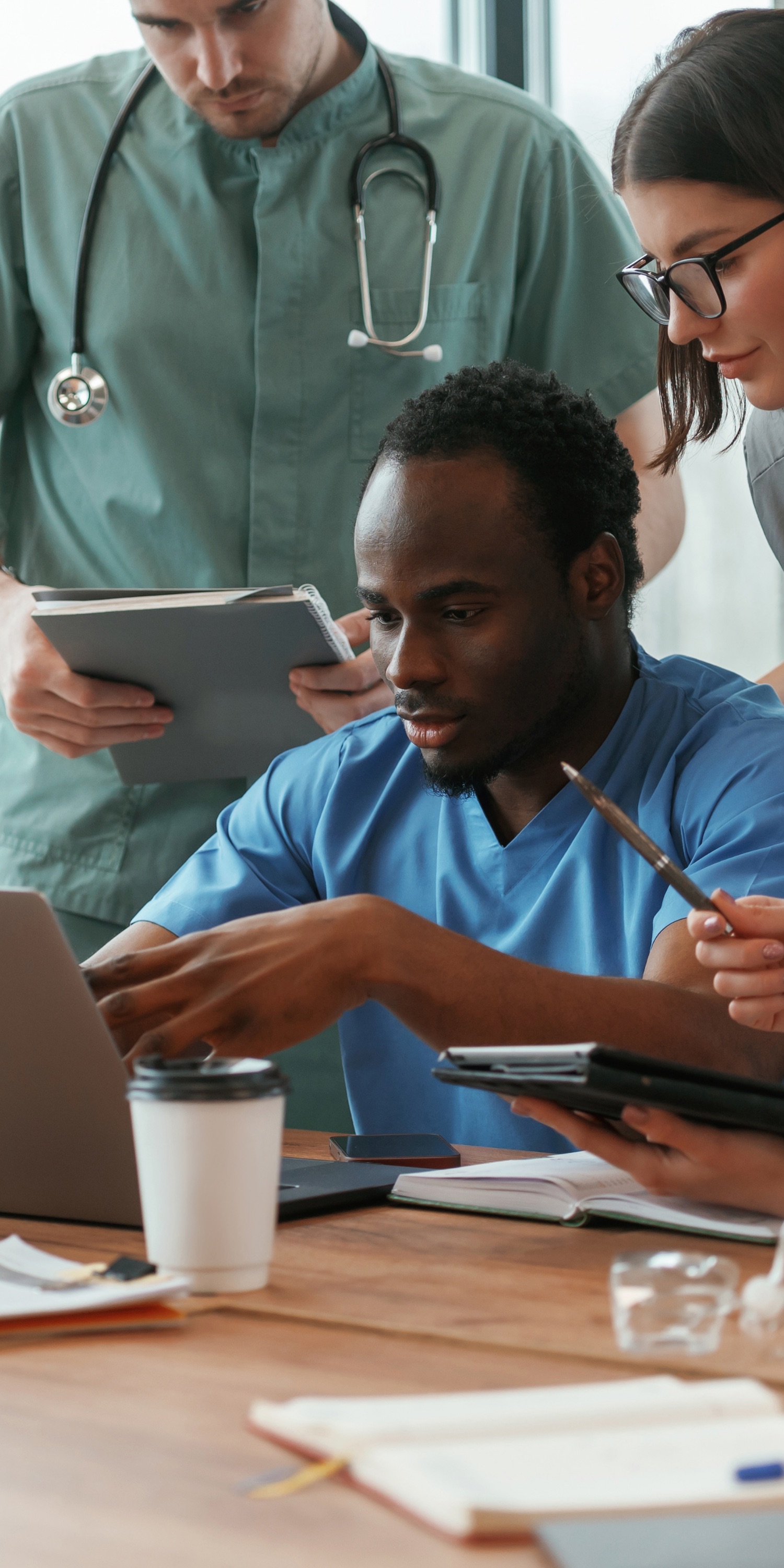 Three medical professionals collaborating while reviewing information on a laptop and taking notes at a wooden table.