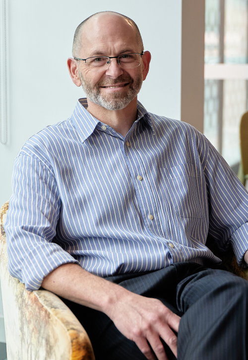 Dr Alisdair Daws posed smiling sitting on a chair with a blue and white striped shirt